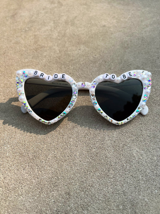 Heart-shaped white sunglasses with bride to be beads written at the top of the frame of the glasses. These bride sunglasses also have pearl and rhinestone accents around the frames. The lense of these bride sunglasses is black UV protected